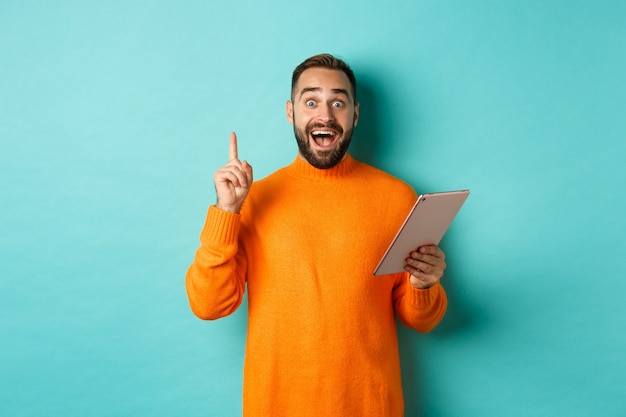 Excited adult man using digital tablet and having an idea, raising finger eureka sign, standing