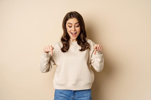 Excited adult woman pointing fingers down and smiling, looking at banner below, showing advertisement, found smth, standing over beige background.