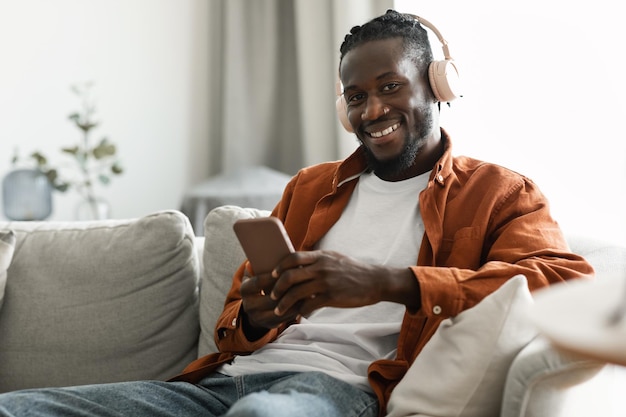 Excited african american man using smartphone while resting on couch at home wearing wireless headset and smiling