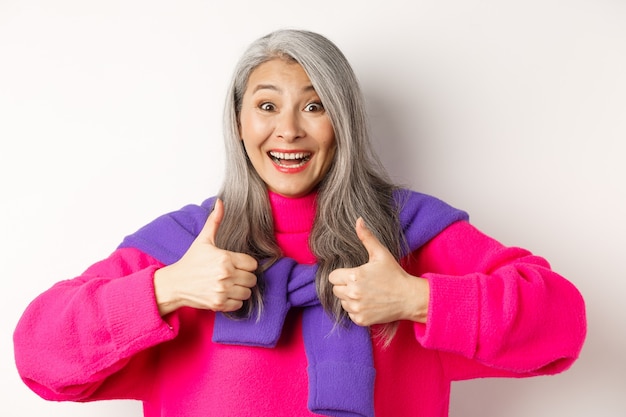 Excited and amazed asian elderly woman showing thumbs-up in approval, smiling happy at camera, praising something awesome, white background.