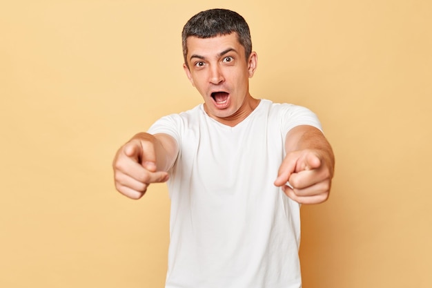 Excited amazed man wearing white casual tshirt standing isolated over beige background pointing to camera with excitement being astonished choosing you