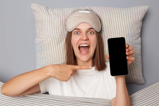 Excited amazed positive woman in white Tshirt and sleeping eye mask lie in bed on pillow under blanket isolated on gray background pointing at smartphone with empty display