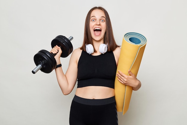 Excited amazed woman holding rolled fitness mat dressed in activewear isolated over gray background doing workout with barbel screaming with positive emotions