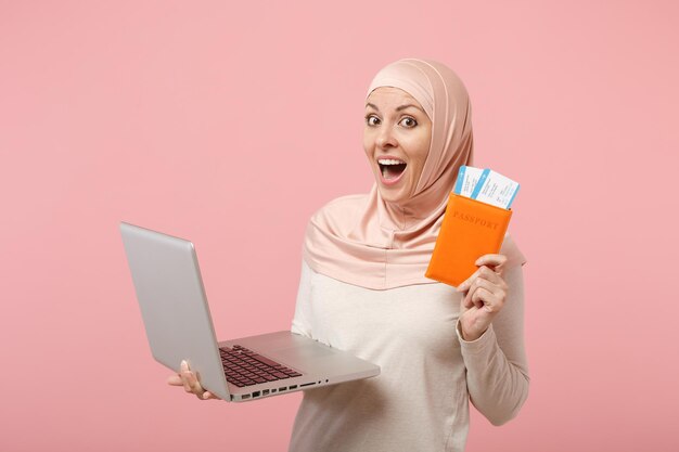 Excited arabian muslim woman in hijab light clothes posing isolated on pink background. People religious Islam lifestyle concept. Mock up copy space. Holding laptop pc computer, passport and ticket.