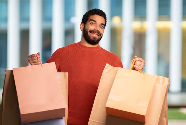 Excited arabic guy showing paper shopping bags posing standing outside