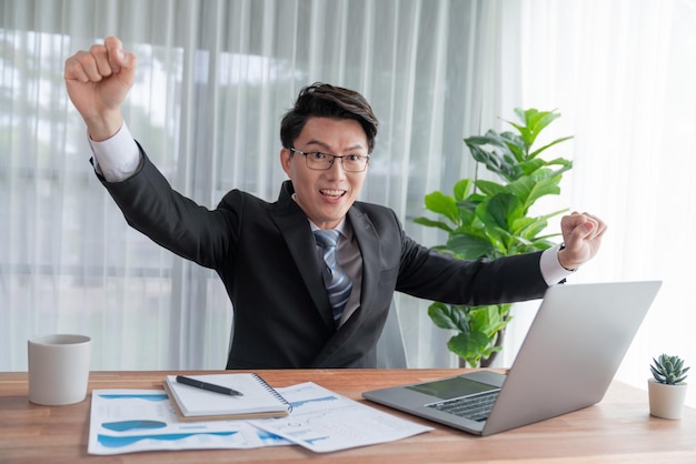 Excited Asian businessman celebrates success at office desk Jubilant