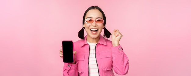 Excited asian girl laughs and smiles shows mobile phone screen smartphone application standing over pink background