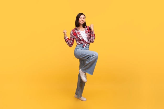 Excited asian lady celebrating success and jumping in air happy having fun over yellow studio background full length