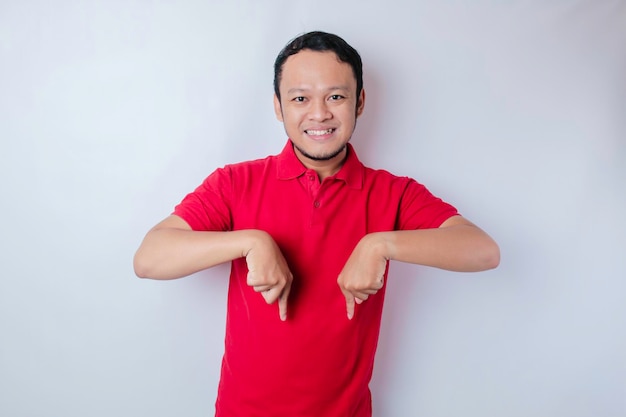 Excited Asian man wearing red tshirt pointing at the copy space below him isolated by white background