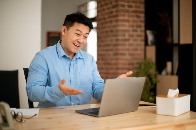 Excited asian mature man using modern laptop video calling and gesturing at webcamera sitting at workplace at home