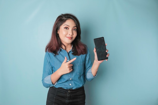 Excited Asian woman wearing blue shirt pointing at the copy space on her smartphone isolated by blue background