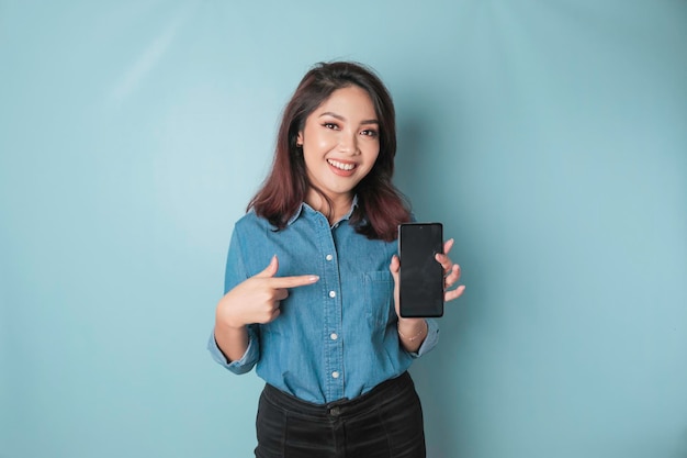 Excited Asian woman wearing blue shirt pointing at the copy space on her smartphone isolated by blue background