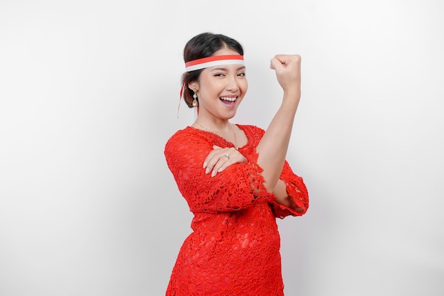 Photo excited asian woman wearing a red kebaya and headband showing strong gesture by lifting her arms and muscles smiling proudly indonesia's independence day concept