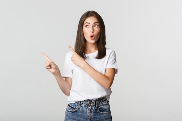 Excited attractive brunette girl in white t-shirt pointing fingers upper left corner, looking curious.