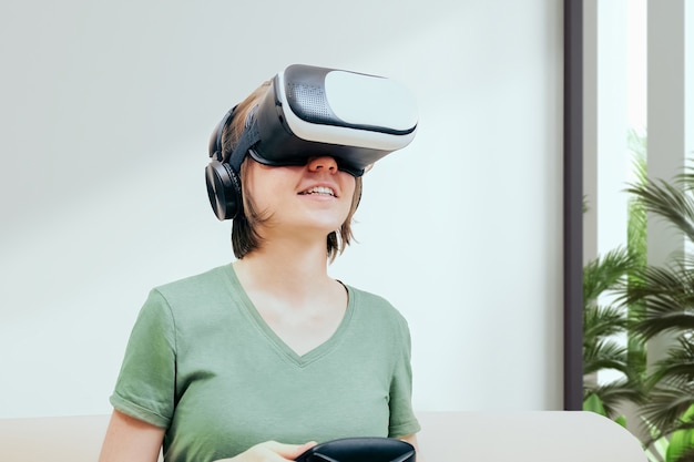 Excited attractive woman gaming with virtual reality headset or goggles and sitting on the couch, holding gamepad controller and playing video game. Virtual reality concept, women in gaming concept