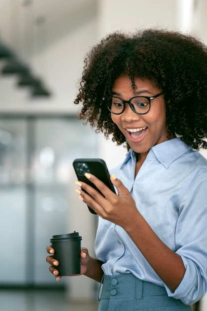 Excited balck woman using mobile phone while relaxing during work standing at office