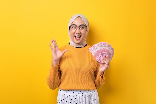 Excited beautiful Asian girl wearing hijab holding money banknotes and celebrating success isolated on yellow background