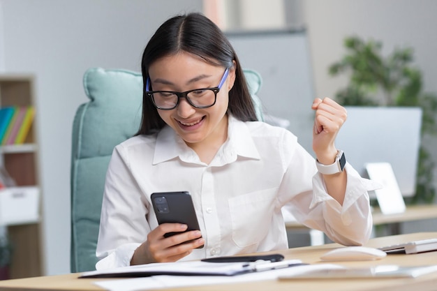 Excited beautiful young asian business woman in glasses win after achievement reading smart phone