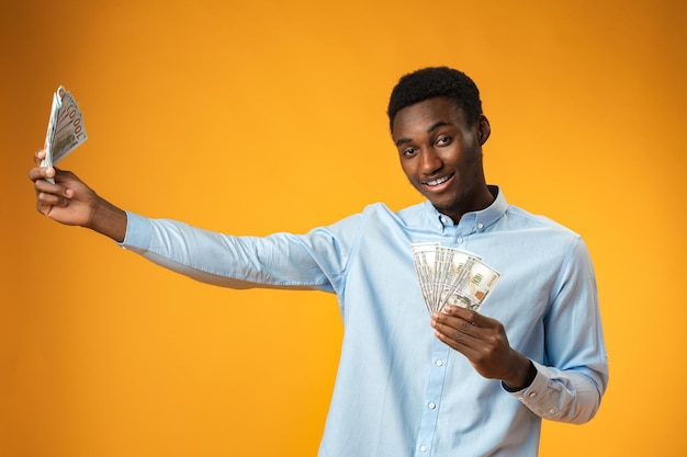 Excited black guy holding stack of dollars in yellow studio