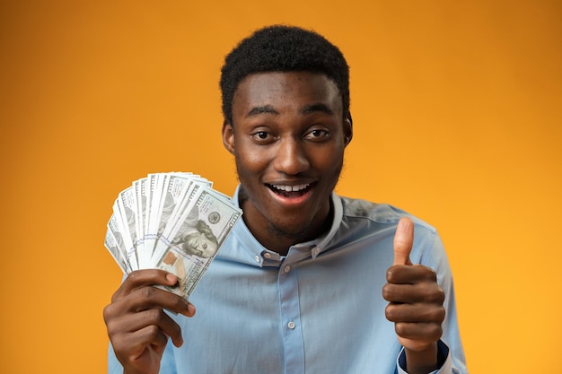 Excited black guy holding stack of dollars in yellow studio