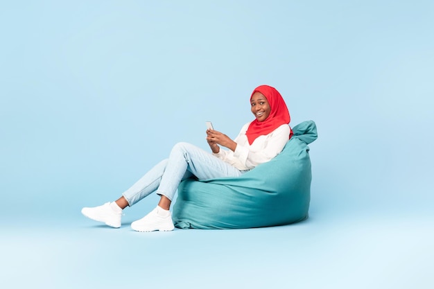 Excited black muslim woman in hijab using smartphone and smiling to camera sitting in beanbag chair blue background