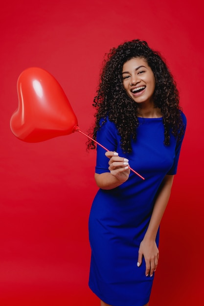 Excited black woman with heart shaped balloon wearing blue dress on red wall