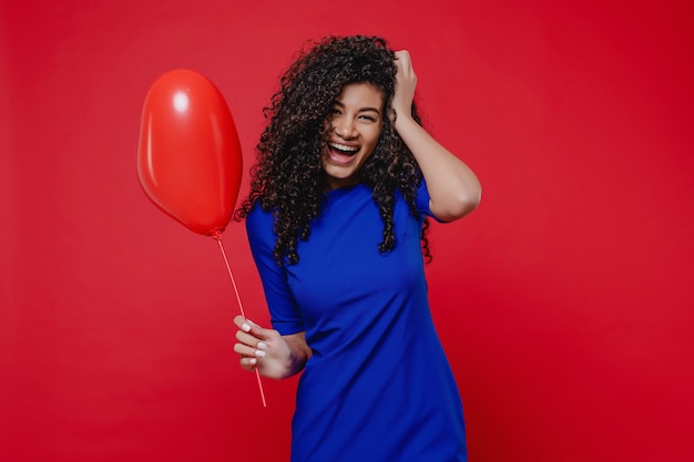 Excited black woman with heart shaped balloon wearing blue dress on red wall