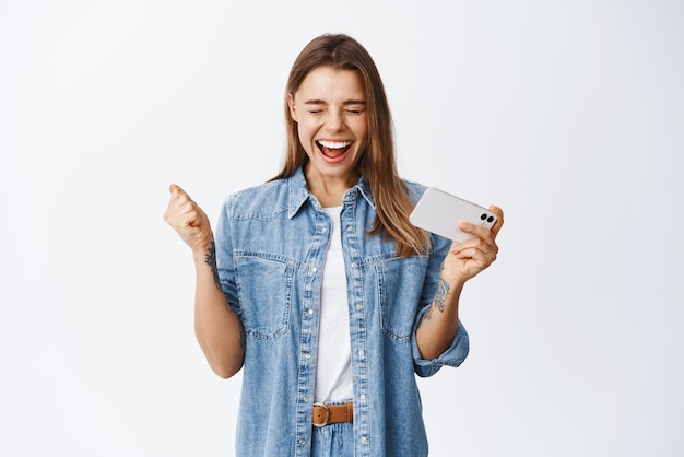 Excited blond woman screaming with happiness holding mobile phone and winning on video game on smartphone cheering and shouting white background