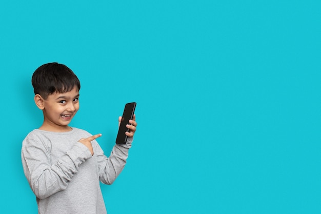 Excited boy smiling and poiting at screen on smartphone. on plain blue background with copy space