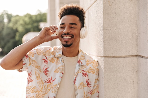 Excited brunet bearded man in floral shirt listens to music in headphones and smiles sincerely with closed eyes outside