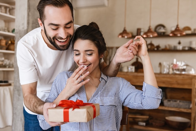 excited brunette couple man and woman having breakfast in apartment while sitting at table with present box