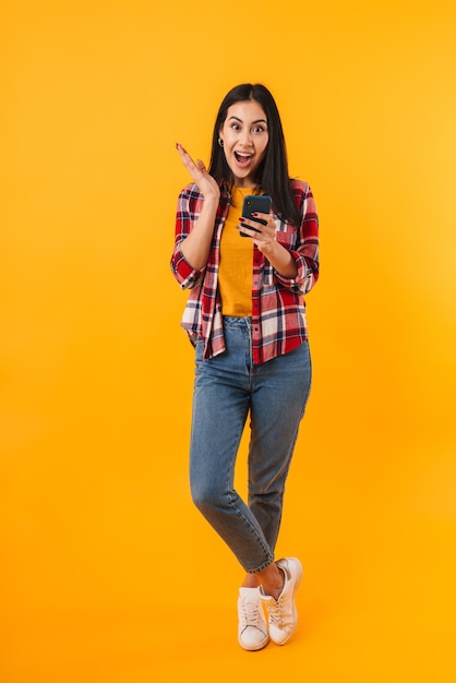 excited brunette woman expressing surprise while using cellphone isolated over yellow wall