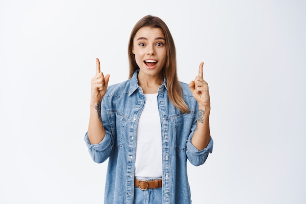 Excited caucasian blond woman gasping amazed, pointing fingers up and showing advertisement, promo deal, standing against white wall