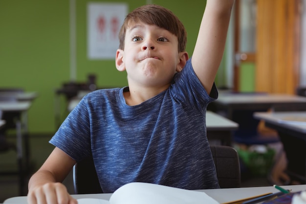 Excited caucasian schoolboy in classroom sitting at desk and raising hand