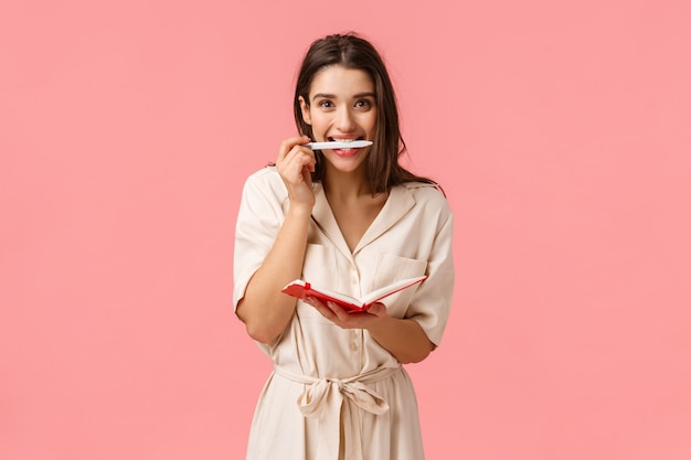 Excited and cheerful young girl stydying new language, smiling holding pen in teeth, writing in cute red notebook, standing pink wall, creating personal schedule, taking notes