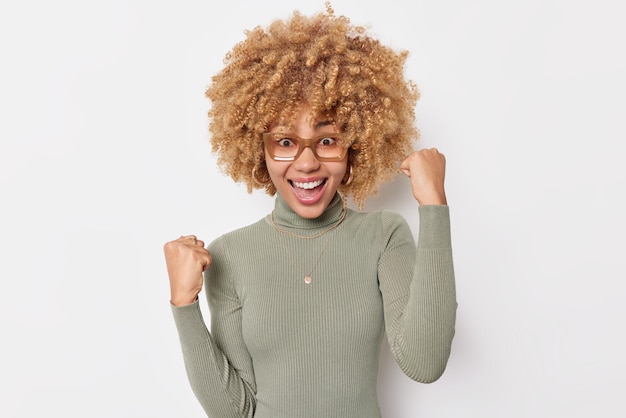 Excited cheerful young woman with curly hair celebrates achievements makes fist pumps has glad expression wears transparent glasses and turtleneck isolated over white background wins something