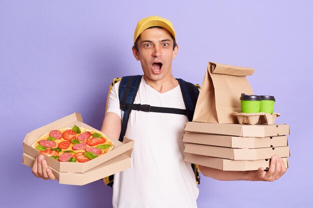 Excited courier man wearing t shirt and cap holding pizza boxes and takeaway coffee in hands looking at camera with open mouth being surprised posing isolated over purple background