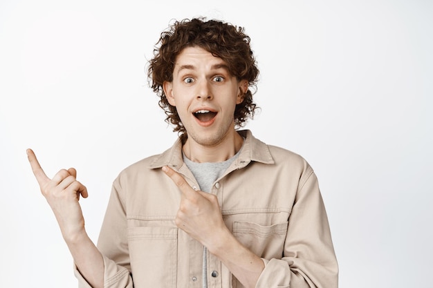 Excited curly guy pointing fingers at upper left corner looking amazed showing smth interesting standing over white background