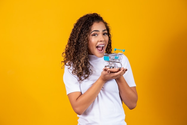 Excited curly hair woman on shopping concept. Young woman with a miniature cart. E-commerce and business. Shopping cart. Buyer woman. Yellow background.