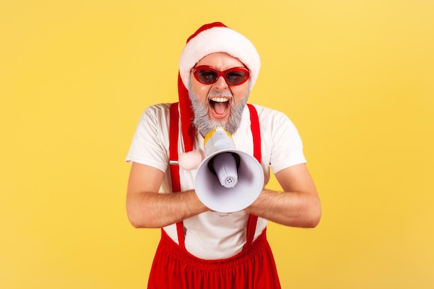 Excited emotional gray bearded man in santa claus costume screaming at loudspeaker, congratulates with holidays, fancy party. Indoor studio shot isolated on yellow background