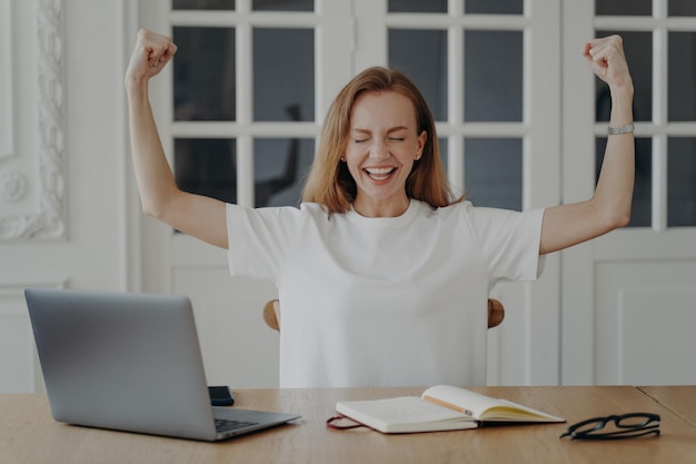 Excited female employee show biceps celebrate good news at desk with laptop Success achievement