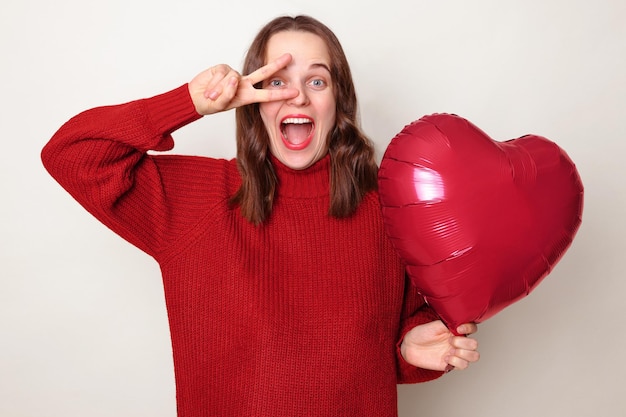 Excited festive woman with brown hair wearing red jumper making victory sign in front her eyes having fun standing isolated over gray background with heart shaped balloon