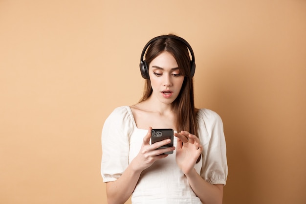 Excited girl listen music in headphones and looking at smartphone amazed, reading awesome news, found cool playlist, standing on beige.