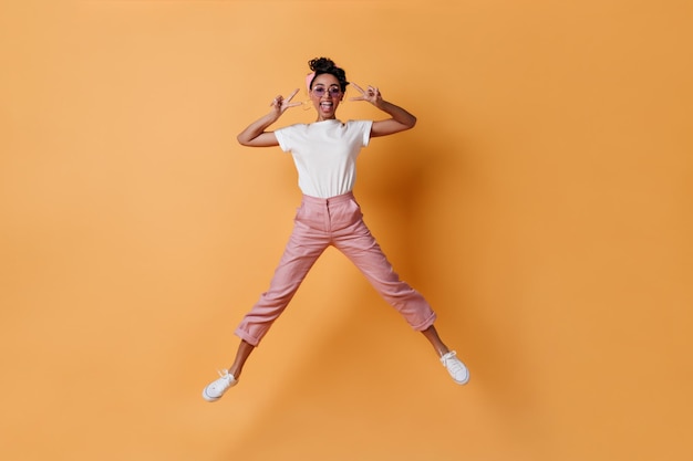 Excited girl in pink pants jumping on yellow background Full length view of mixed race lady showing peace signs