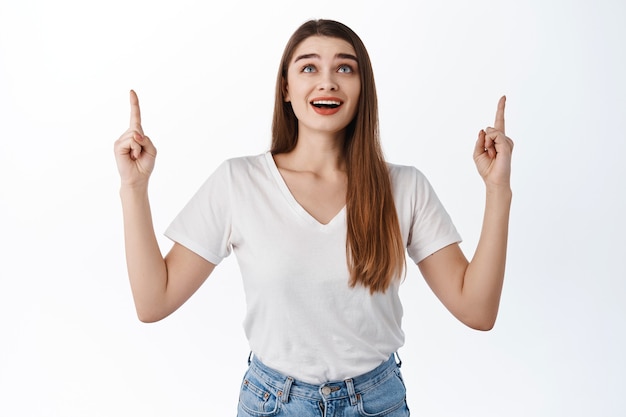 Excited girl shows advertisement on top, looking up with fascinated smiling face, checking out awesome promo deal, staring at banner logo, standing over white wall