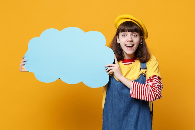 Excited girl teenager in french beret, denim sundress hold blue empty blank Say cloud, speech bubble isolated on yellow wall background. People sincere emotions, lifestyle concept. Mock up copy space.