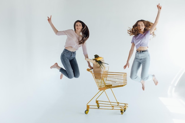 excited girls with shopping bag sitting in shopping cart, isolated on white