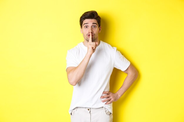 Excited guy planning surprise, shushing at front, press finger to lip in hush sign, standing over yellow wall