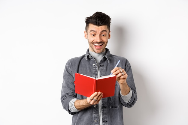 Excited guy writing in planner and smiling, writing down ideas in journal or diary, standing on white background.