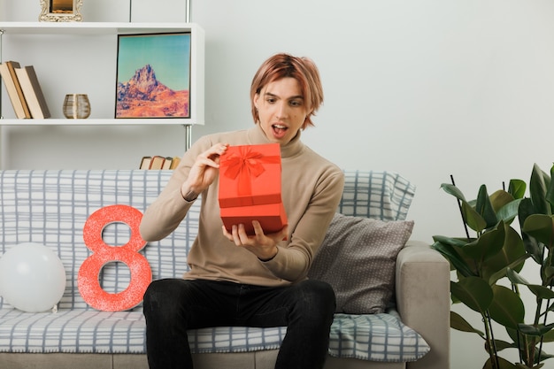 Excited handsome guy on happy women day holding and looking at present sitting on sofa in living room